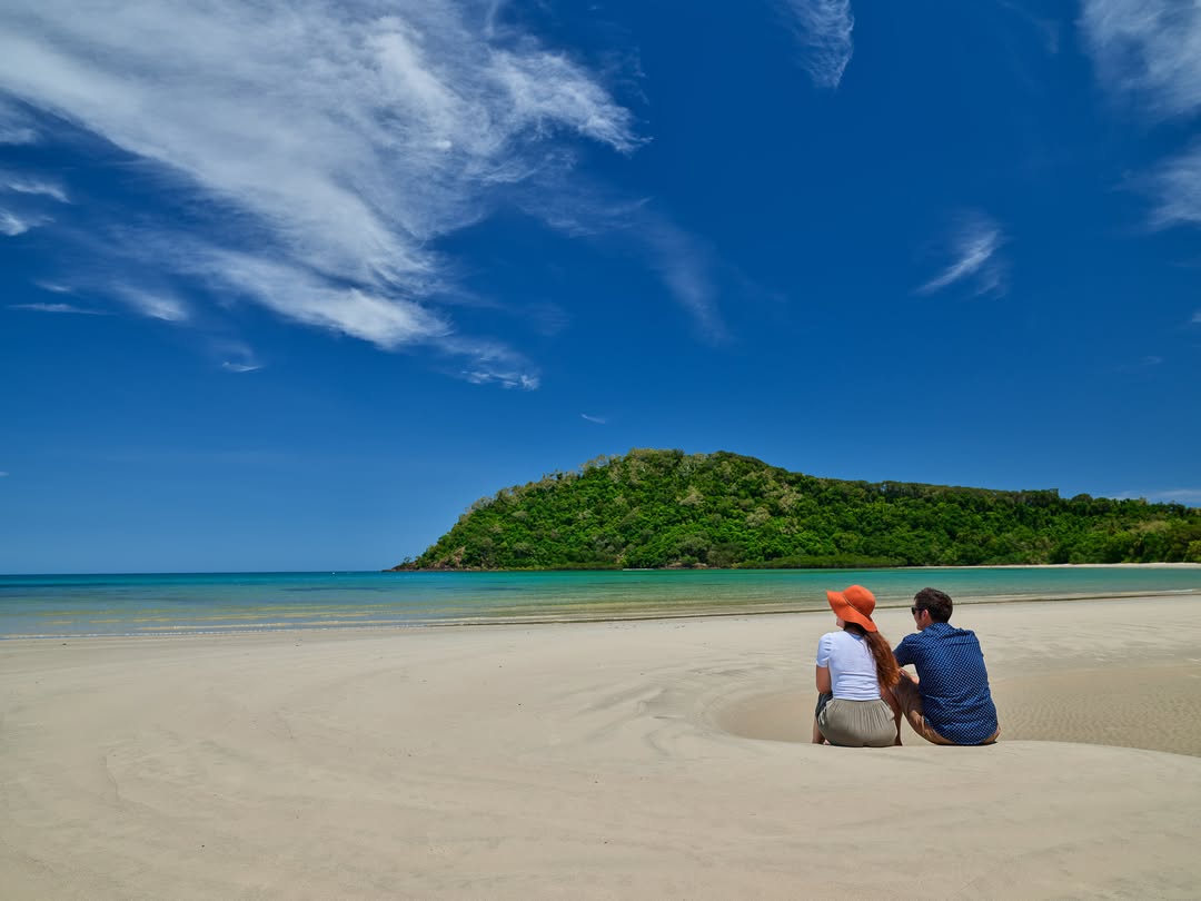 Couple relaxing on the sand during a Cape Tribulation day trip with Down Under Tours, overlooking turquoise water, tropical coastline, and lush rainforest-covered hills beneath a bright blue sky.v