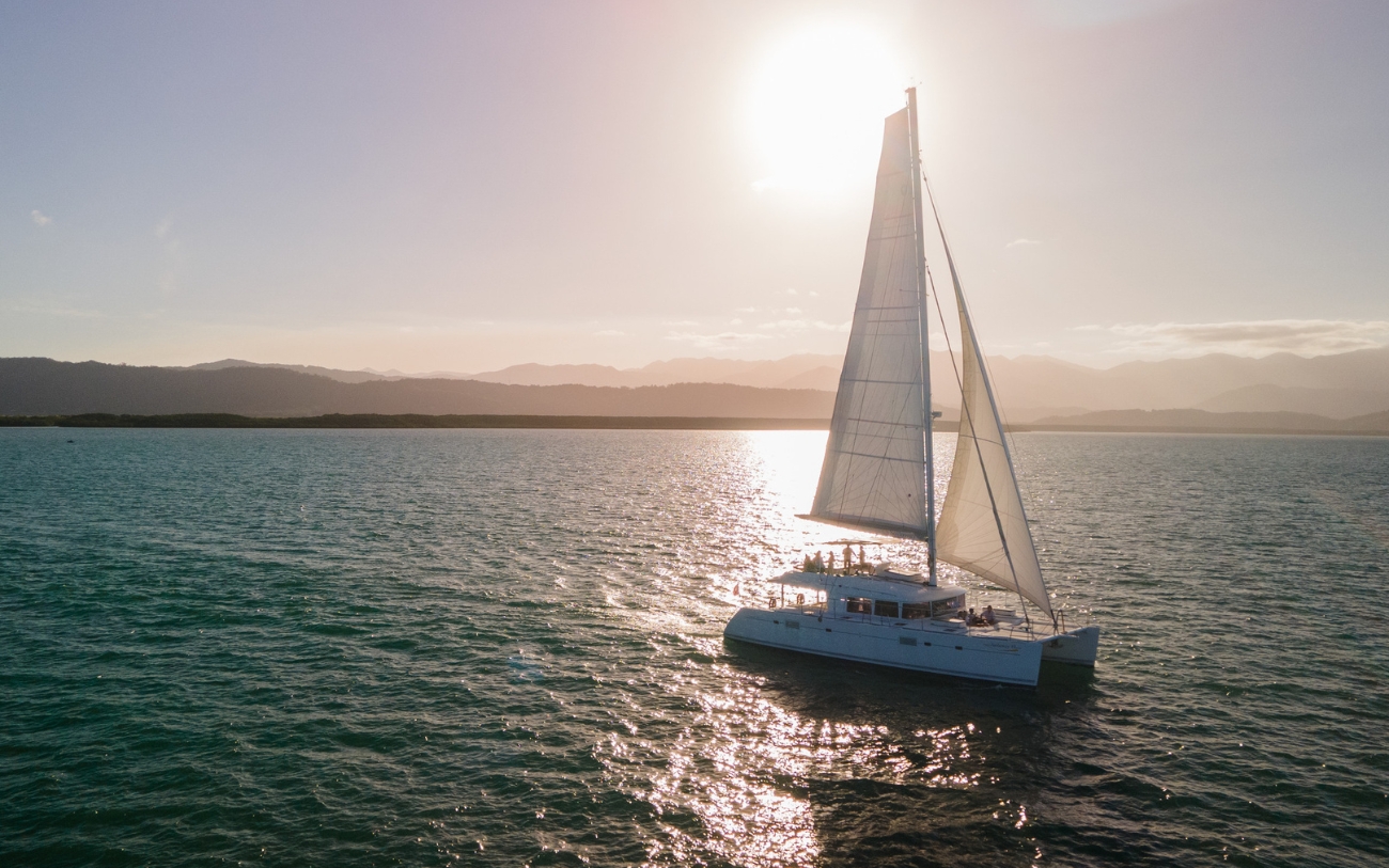 Sailaway catamaran cruising across calm ocean waters during an afternoon sunset tour near Port Douglas, with golden sunlight reflecting across the sea and mountain ranges in the distance.