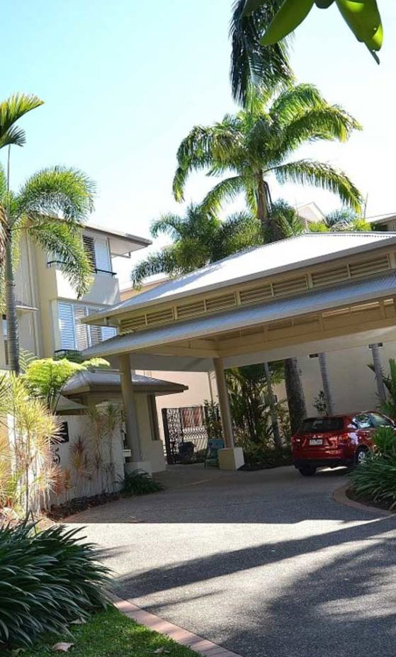 Cayman Villas entrance in Port Douglas featuring tropical landscaping, palm trees, and a red car parked in the driveway.