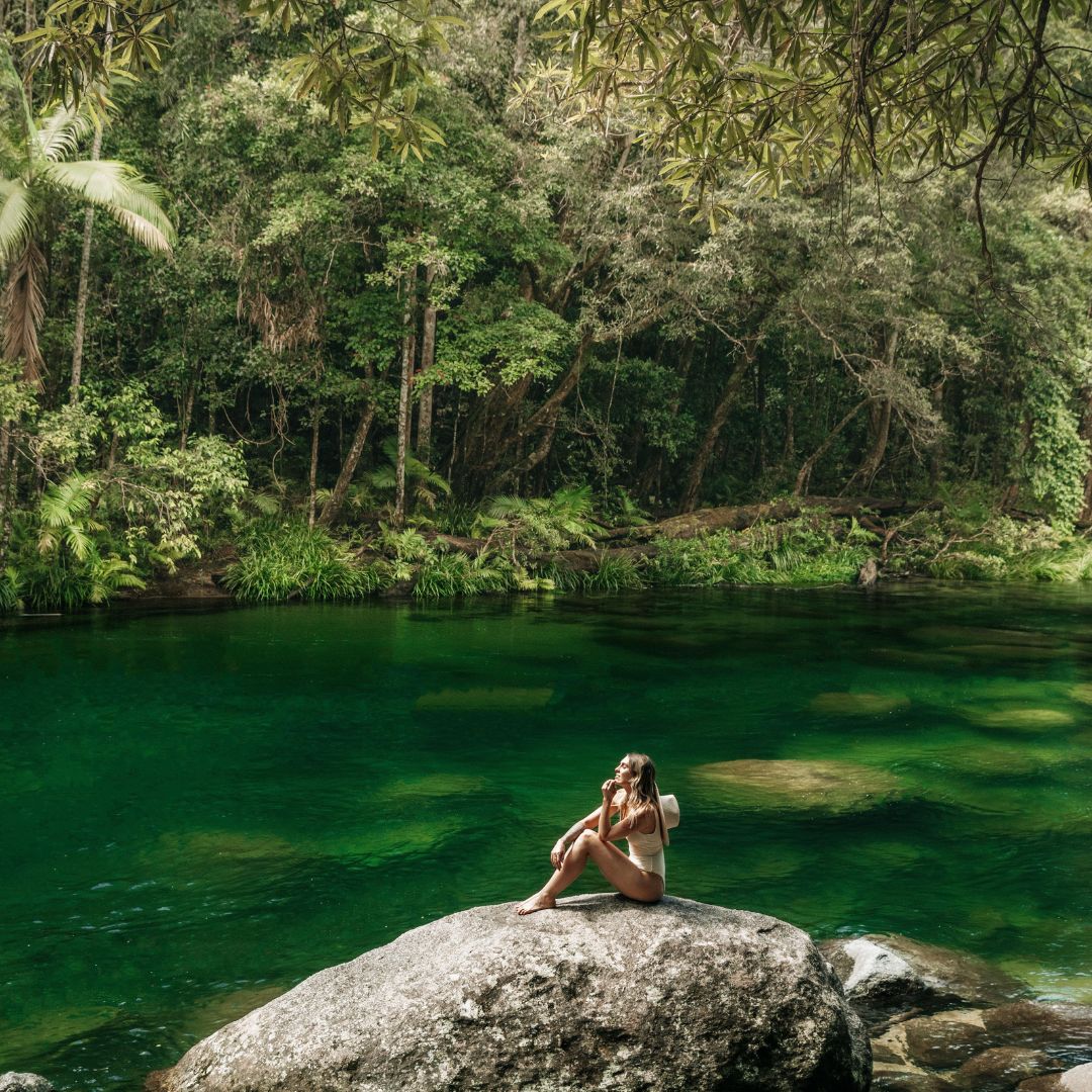 Woman sitting on a large rock beside the emerald waters of Mossman Gorge surrounded by lush tropical rainforest near Port Douglas.