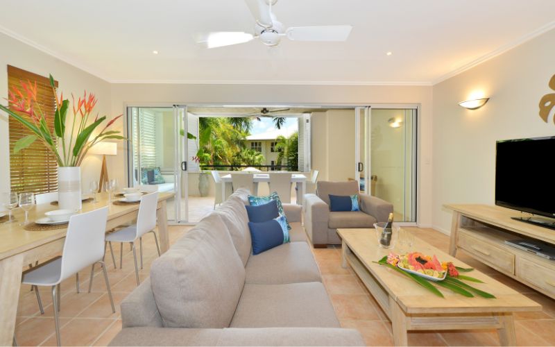 Bright, modern living area at Cayman Villas Port Douglas featuring a lounge, dining space, tropical décor, and open sliding doors leading to a private balcony surrounded by palm trees.