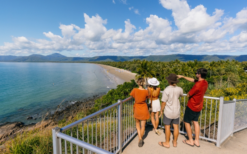 Group of visitors enjoying panoramic views from Flagstaff Hill Lookout in Port Douglas, overlooking Four Mile Beach, tropical coastline, and distant mountain ranges under a bright blue sky.