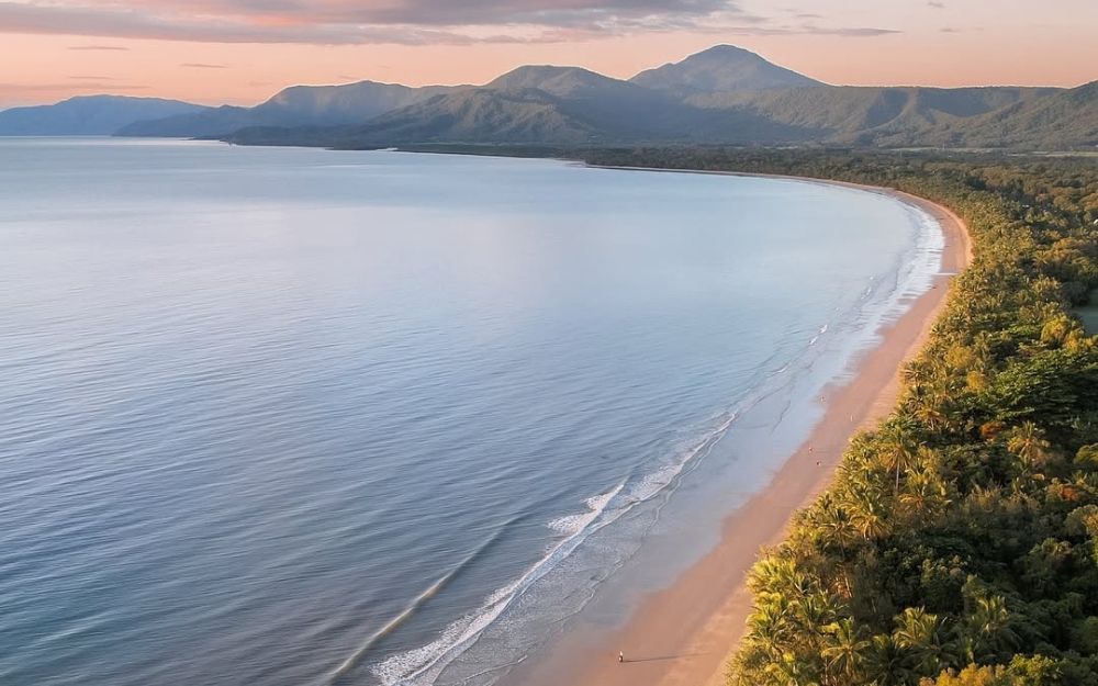 Aerial view of Four Mile Beach with golden sand stretching along the coastline, framed by tropical rainforest and mountains at sunrise.