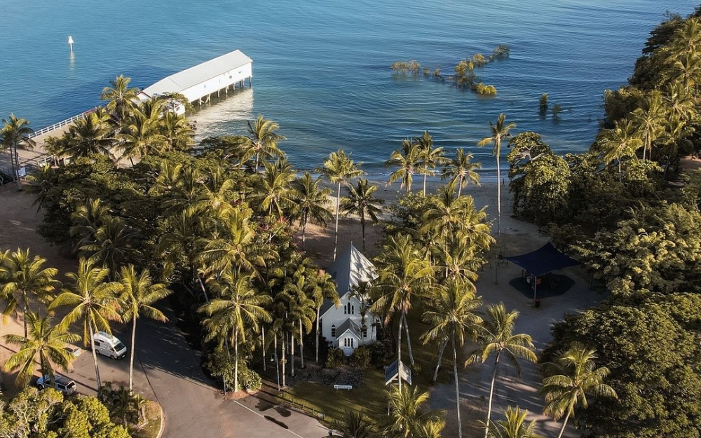 Aerial view of St Mary's by the Sea chapel and the historic Sugar Wharf in Port Douglas, surrounded by tropical palm trees and overlooking the calm waters of Dicksons Inlet.