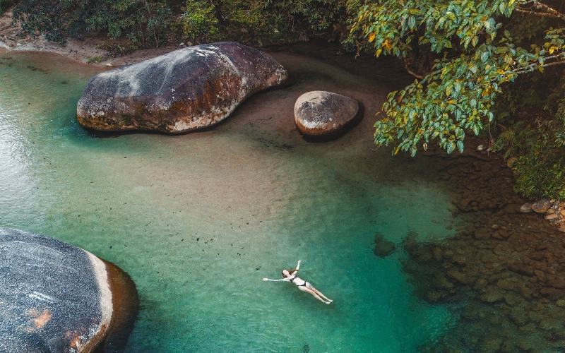 Swimmer floating in the clear turquoise water at Babinda Boulders, surrounded by large granite boulders and tropical rainforest.