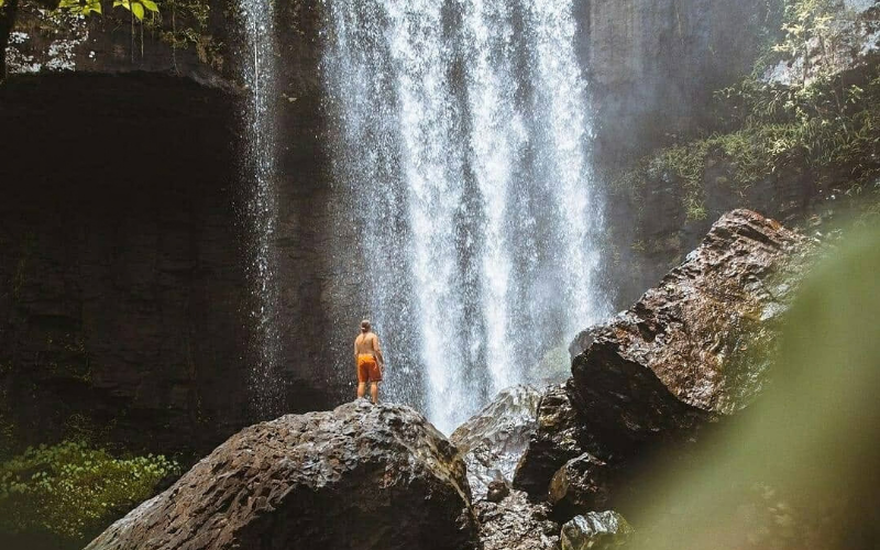 Person standing on large rocks beneath the powerful Zillie Falls in the lush Atherton Tablelands rainforest.